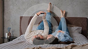 Caucasian happy couple lying upside down together on comfort bed relax with mobile phone scrolling social network buying