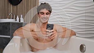 Caucasian happy smiling man in foam bath washing relaxing in foamy bathe using mobile phone scrolling smartphone gadget