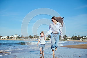 Caucasian happy young mother and daughter smiling while running on the beach, leaving footsteps on the wet sand