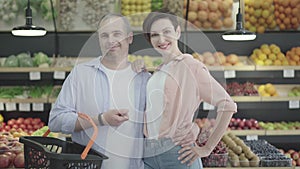 Caucasian husband and wife hugging and smiling at camera in grocery. Portrait of happy young beautiful brunette woman