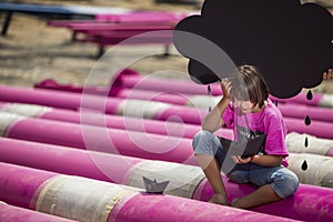 Caucasian kid sits on a pink pipes with black paper boats and clouds at the construction site