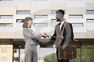 Caucasian lady welcomes African American male colleague, shaking hands near office building