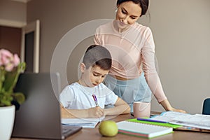 Caucasian little teen boy writing homework in copybook and mother standing by. Checking.