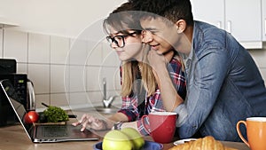 Caucasian looking girl and her mulatto boyfriend both in their 20`s using laptop, surfing the web in the kitchen at home