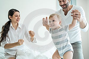 Caucasian loving parents play with baby boy child on bed in bedroom. Happy family, attractive beautiful young couple dad and mom