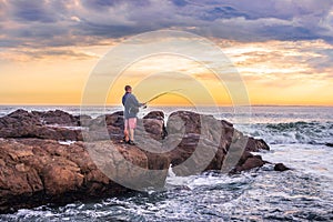 Caucasian male on the beach at sunset fishing from the rocks on the shoreline, with Table Mountain and Cape Town coastline