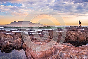 Caucasian male on the beach at sunset fishing from the rocks on the shoreline, with Table Mountain and Cape Town coastline