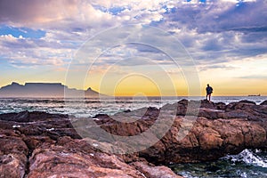 Caucasian male on the beach at sunset fishing from the rocks on the shoreline, with Table Mountain and Cape Town coastline
