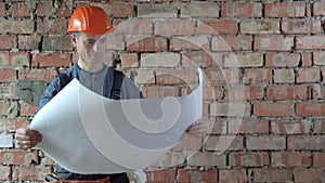 A Caucasian male builder stands against a background of a red brick wall dressed in an orange hard hat and a uniform