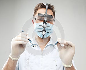 Caucasian male doctor in a surgical mask and binocular loupes holding dental instruments at the grey background