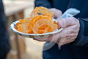 Caucasian male hand holds a plate of Jalebi tradtional Indian fried sweet dessert at a festival, in selective focus photo