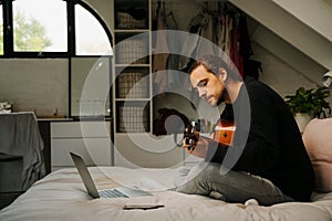 Caucasian male learning how to play the guitar, watching an online lesson at home on the bed