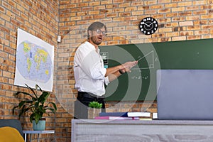 Caucasian male maths teacher standing by blackboard giving online lesson using laptop