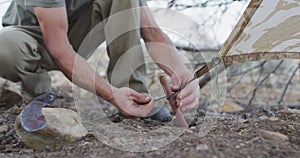 Caucasian male survivalist attaching tent to stick peg at camp in wilderness