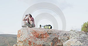 Caucasian male survivalist using binoculars, sitting on mountain peak in wilderness