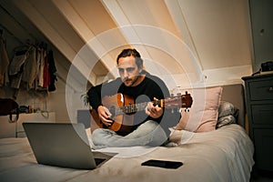 Caucasian male teen playing guitar learning songs off internet on laptop relaxing in comfortable bedroom