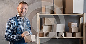 Caucasian male worker dressed in a blue overalls stands near a shelf with boxes makes a pen marks on the box. Copy space