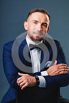 Caucasian man of 30-35 years old in business classic suit sits on chair and looks straight into camera. Studio portrait
