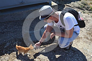 Caucasian man with a backpack crouched down and holding an action camera in his outstretched hand. Red kitten sniffs the camera