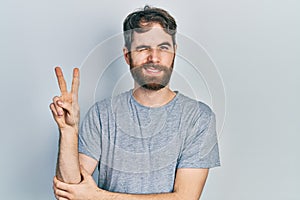 Caucasian man with beard wearing casual grey t shirt smiling with happy face winking at the camera doing victory sign