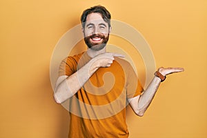 Caucasian man with beard wearing casual yellow t shirt amazed and smiling to the camera while presenting with hand and pointing