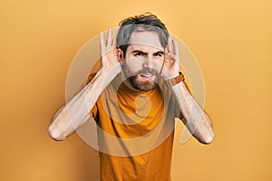 Caucasian man with beard wearing casual yellow t shirt trying to hear both hands on ear gesture, curious for gossip