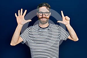 Caucasian man with beard wearing striped t shirt and glasses showing and pointing up with fingers number seven while smiling