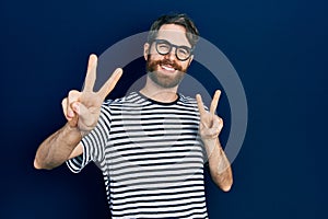 Caucasian man with beard wearing striped t shirt and glasses smiling looking to the camera showing fingers doing victory sign