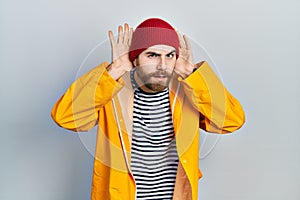 Caucasian man with beard wearing yellow raincoat trying to hear both hands on ear gesture, curious for gossip