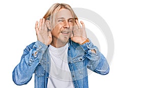 Caucasian man with blond long hair wearing casual denim jacket trying to hear both hands on ear gesture, curious for gossip