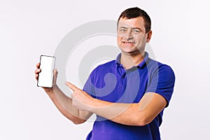 Caucasian man in blue t-shirt looks happy at the camera and shows his forefinger at the mocks up smartphone screen. Gray photo