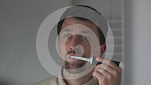 Caucasian man brushes his teeth with an electric toothbrush in the bathroom, view of the reflection through the mirror