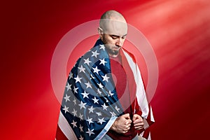 A Caucasian man covered with an American flag stands in a humble and proud pose. Red background. Copy space. Concept of memorial