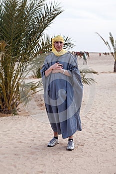 Caucasian man dressed in traditional dishdasha and yellow headscarf standing in desert against palm tree, an European tourist photo