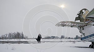 A Caucasian man with a guitar case walks next to an old Soviet-made AN 2 aircraft.