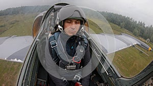 Caucasian man in a helmet - pilot of gray combat fighter plane boring and flying over green field and forest in cloudy sky