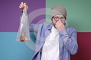 Caucasian man holds a stinky, spoiled fish in a transparent package and pinches nose with his fingers due to an