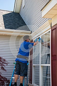 A Caucasian man on a ladder wearing blue latex gloves and listening to ear buds washing a window with a brush
