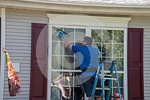 A Caucasian man on a ladder wearing blue latex gloves and listening to ear buds washing a window with a squeegee