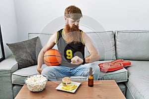 Caucasian man with long beard holding basketball ball cheering tv game checking the time on wrist watch, relaxed and confident