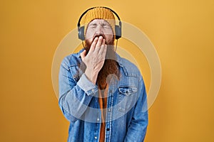 Caucasian man with long beard listening to music using headphones bored yawning tired covering mouth with hand