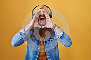 Caucasian man with long beard listening to music using headphones shouting angry out loud with hands over mouth