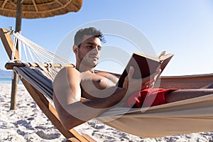 Caucasian man lying on a hammock and reading a book at the beach.