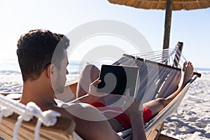 Caucasian man lying on a hammock and using a digital tablet at the beach.