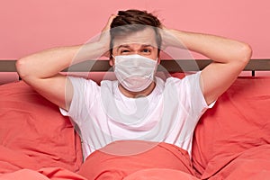 Caucasian man in medical mask covering ear having a panic attack resting on bed