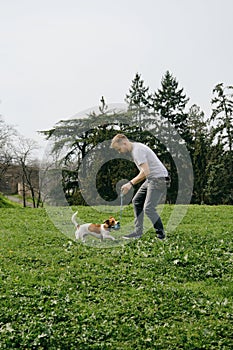 Caucasian man plays toy rope tug-of-war with a dog on green grass. An active spring walk in park of male owner and jack