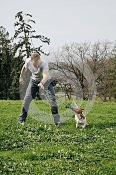 Caucasian man plays toy rope tug-of-war with a dog on green grass. An active spring walk in park of male owner and jack