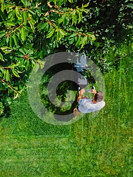 Caucasian man pushing lawn mower for cutting green grass in garden with sunlight at summer season. Aerial view. Housework and