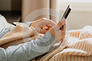 A caucasian man relaxing, using smart phone lying in bed under throw at home