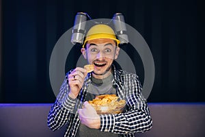 Caucasian man rests at home and watches TV shows or sports news on TV screen. Man with beer helmet on the head eats